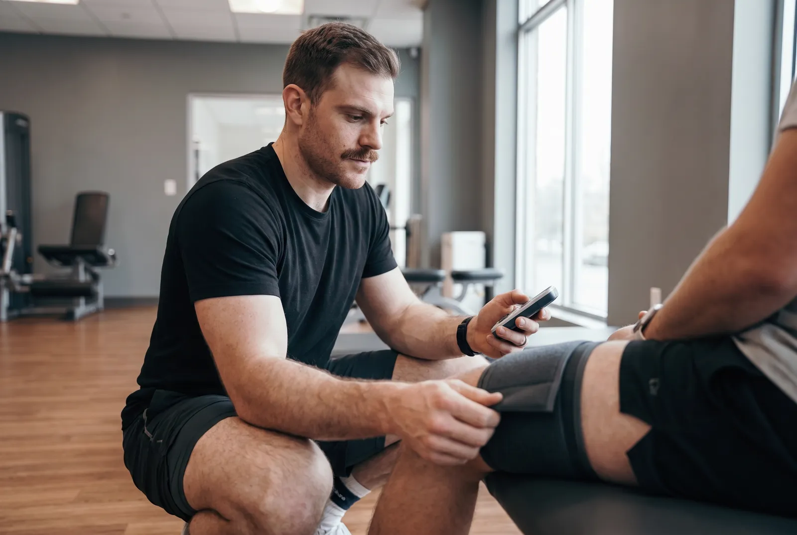 A physical therapist adjusting a cuff on a patient's thigh while checking a handheld device